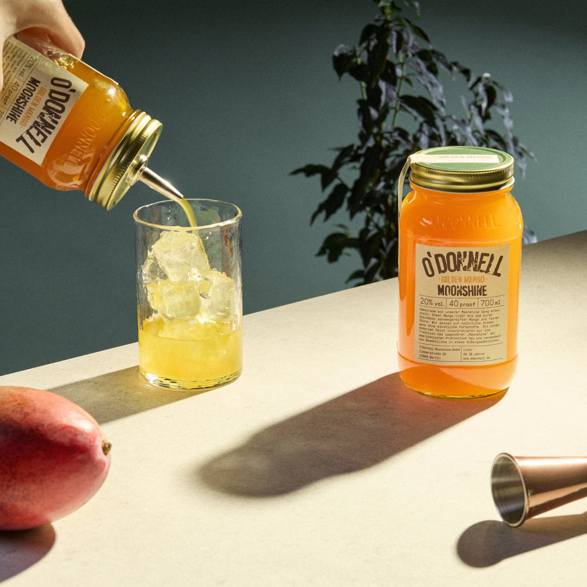Auf der rechten Seite ein Glas Golden Mango Likör, auf der linken Seite ein Glas voller Eis und eine Person, die Mango Likör aus einem anderen Glas einschenkt.