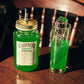 Ein großes, grünes Likörglas der Sorte „Waldmeister“ mit Ausgießer steht neben einem hohen Drink mit Eisstange auf Holz.
