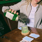 Eine Person gießt grünen „Waldmeister“ Likör aus einem großen O'Donnell Glas in ein strukturiertes Cocktailglas auf einem Holztisch.