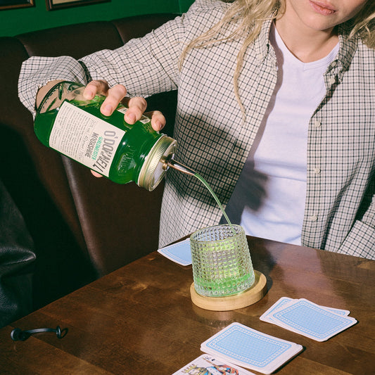 Eine Person gießt grünen „Waldmeister“ Likör aus einem großen O'Donnell Glas in ein strukturiertes Cocktailglas auf einem Holztisch.